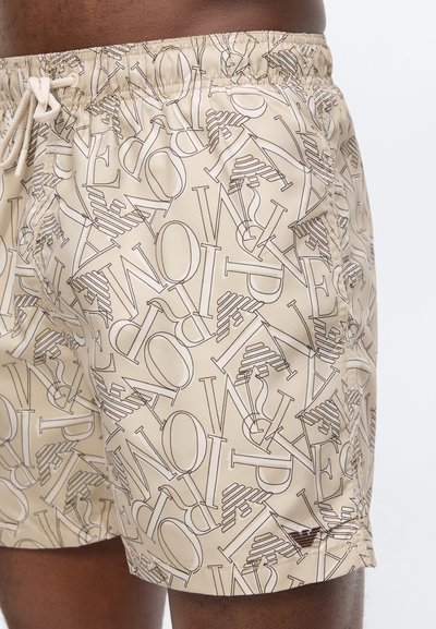 Μπεζ μαγιό σορτς με ελαστική μέση και κορδόνι, με διάσπαρτα μεγάλα περιγραμμένα γράμματα και ένα μικρό σκουρόχρωμο λογότυπο κοντά στο τελείωμα.