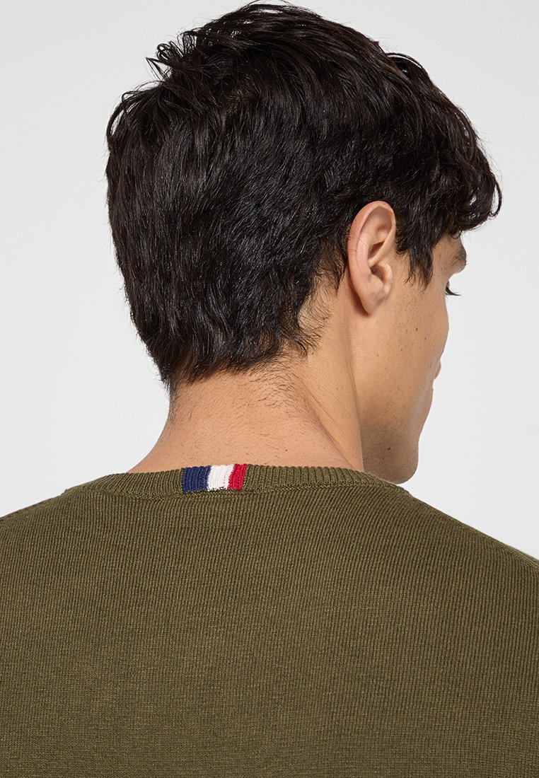 Uomo con capelli corti scuri che indossa un maglione a maglia verde oliva con un piccolo dettaglio a strisce blu, bianco e rosso sul colletto posteriore.