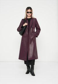 Burgundy faux leather trench coat with double-breasted buttons, side pockets, and a relaxed fit, paired with black knee-high boots and a handbag.