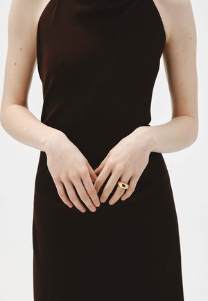 Vestido negro sin mangas con textura suave, combinado con un anillo dorado que presenta un diseño de corte ovalado en la parte frontal. Las manos están suavemente entrelazadas.