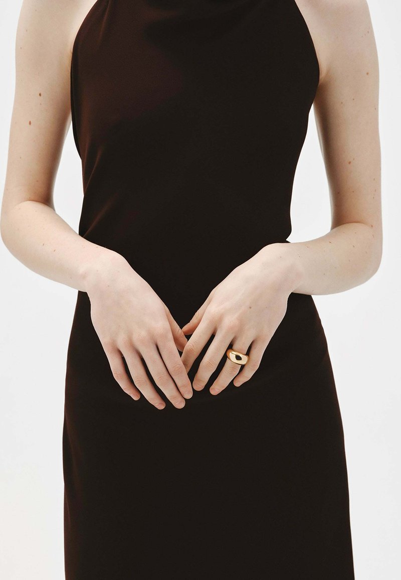 Vestido negro sin mangas con textura suave, combinado con un anillo dorado que presenta un diseño de corte ovalado en la parte frontal. Las manos están suavemente entrelazadas.