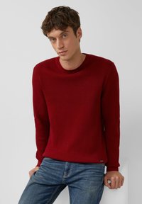 Roter strukturierter Strickpullover mit Rundhalsausschnitt und langen Ärmeln, kombiniert mit blauen Jeans. Verfügt über ein dezentes Markenetikett in der Nähe des Saums.