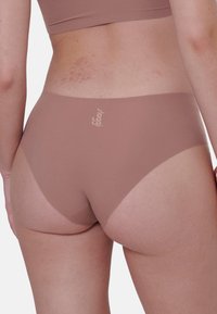 Beige saumatonta alusvaatetusta, jossa on sileä rakenne, keskipitkä leikkaus ja minimaalinen takakattavuus. Sisältää hienovaraisen logon painatuksen vyötäröllä.