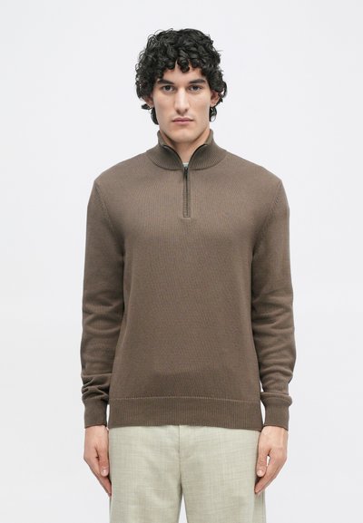 Pull en laine marron à fermeture éclair avec un col haut, texture côtelée et manches longues, porté avec un pantalon clair texturé.