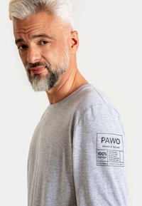 Jasnoszara koszula z długim rękawem, wykonana w 100% z bawełny, z metką na lewym rękawie zawierającą nazwę marki i instrukcje pielęgnacji.