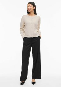 Gaiši bēšs kardigans ar apaļu kakla izgriezumu un pogu aizdari, kombinēts ar plašām melnām biksēm un melnām apaviem ar smailiem purngaliem.