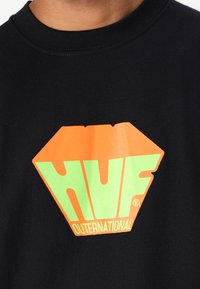 Zwarte katoen t-shirt met een groot diamantvormig logo in oranje en groen, met de tekst "HUF OUTERNATIONAL" aan de onderkant.