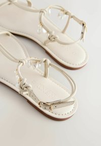 Sandales plates blanches avec brides à la cheville, boucles dorées, détail noué à l'avant, et petites perles transparentes pendantes sur un fond uni.