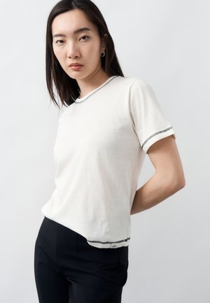 Νεαρή γυναίκα στέκεται με τα χέρια πίσω από την πλάτη, φοράει λευκή κοντομάνικη μπλούζα με μαύρη διακόσμηση ραφής και μαύρο παντελόνι, μπροστά σε απλό φόντο.