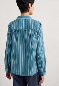Niebieska, paskowana koszula z długim rękawem, z kołnierzykiem, plisowanym tyłem oraz zapięciem na guziki przy mankietach. Miękka tkanina w pionowe białe paski.