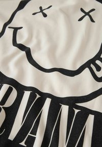 Bomullst-skjorte med et svart smiley-ansikt og ordet "NIRVANA" i fete svarte bokstaver på en kremfarget bakgrunn, med en jevn tekstur.