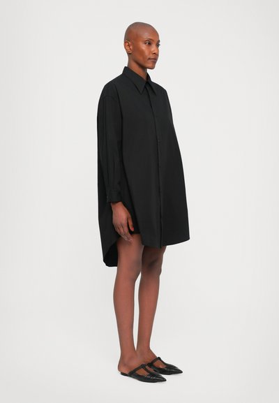 Black oversized shirt dress with a button-down front, long sleeves, and a high-low hem, paired with black slingback shoes. Smooth fabric.