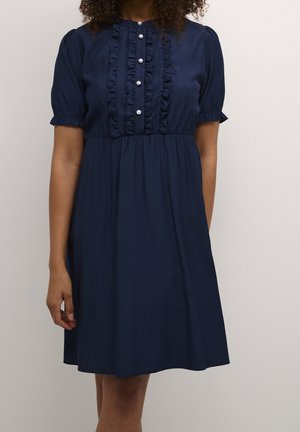 Frau trägt ein marineblaues knielanges Kleid mit kurzen Puffärmeln, gerafften Frontpaneelen und sechs Perlenknöpfen entlang der Brust.