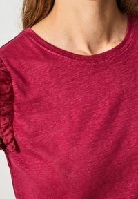 Žena oblečená v texturovaném hluboce červeném tričku s krátkým rukávem, které má zdobení ve formě volánků na rukávech a kulatý výstřih.