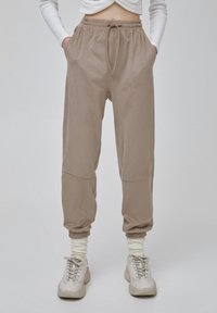 Modellen bär beiga joggerbyxor med dragsko i midjan och sidofickor, i kombination med vita strumpor och ljusa sneakers mot en neutral bakgrund.