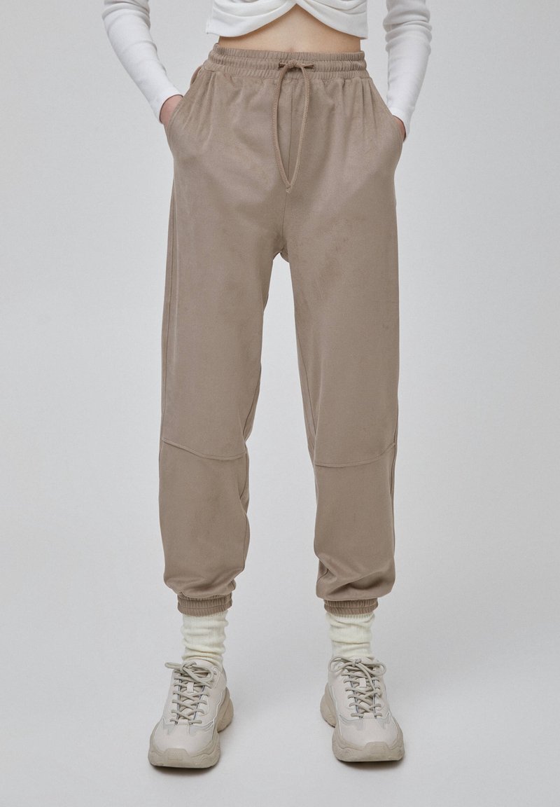 Modellen bär beiga joggerbyxor med dragsko i midjan och sidofickor, i kombination med vita strumpor och ljusa sneakers mot en neutral bakgrund.