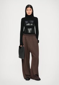 Svart ribbad turtleneck-tröja med glansiga horisontella ränder, matchad med löst sittande bruna byxor och en svart clutch-väska.