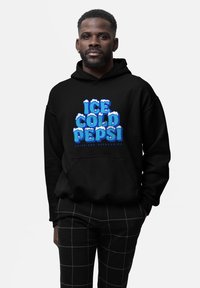 Uomo con capelli corti e barba, che indossa una felpa nera con la scritta "Ice Cold Pepsi" e pantaloni a scacchi neri, in piedi contro uno sfondo bianco.