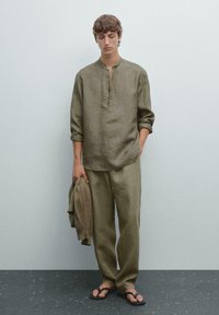 Jonge man in een losse olijfgroene linnen blouse en broek met opgerolde mouwen en zwarte sandalen, die een sweater vasthoudt tegen een plain muur.