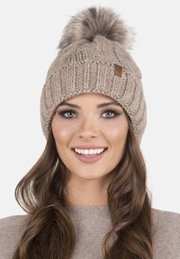 Caciula tricotată chunky, de culoare bej, cu un pompon din blană sintetică în vârf și o etichetă mică din piele, având o textură ribbed pe întreaga suprafață.