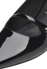 Chaussure en cuir verni noir avec un bout pointu, dotée d'une bride croisée et d'un accent métallique sur le côté. Texture de surface lisse et brillante.