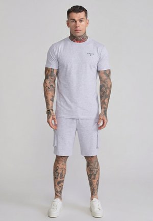 Mężczyzna z tatuażami na szyi, ramionach i nogach noszący jasnoszarą koszulkę i szorty SikSilk oraz białe trampki, stojący na jednolitym tle.