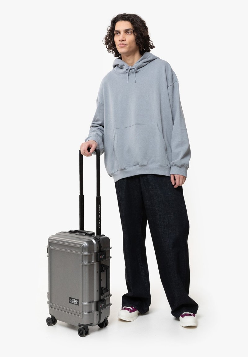 Gray hard-shell suitcase with retractable handle, textured surface, and two wheels. Individual wearing a gray hoodie and dark, loose trousers.