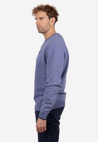 Hombre con cabello rizado, vestido con una sudadera azul de manga larga y pantalones oscuros, de pie de perfil contra un fondo blanco uniforme.