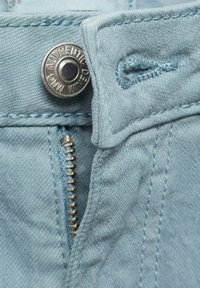 Svetlomodrá denimová tkanina má zips a kovový gombík označený "AUTENTICKÁ DENIMOVINA." Detailný pohľad na švy.