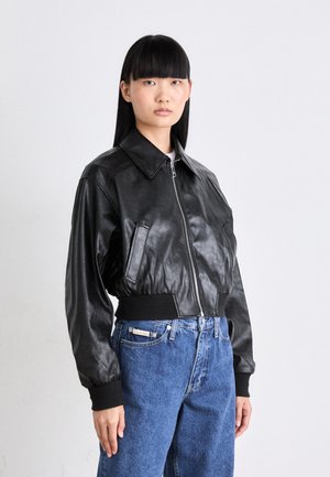 Joven mujer con cabello lacio negro, vistiendo una chaqueta de cuero negra con cierre y jeans azul Calvin Klein frente a un fondo liso.