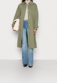 Olijfgroen trenchcoat met grote knopen, een kraag en manchetdetails, gedragen over lichtblauwe wijde jeans en gecombineerd met een crèmekleurige handtas.