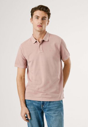 Polo shirt rose clair à manches courtes avec un col et un bouton unique, présentant un petit logo brodé sur la poitrine. Porté avec un jean bleu.