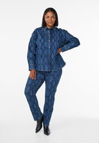 Modrý denimový outfit s hadím vzorem, košilí na knoflíky a odpovídajícími kalhotami. Kalhoty mají rovný střih s mírným elastanem.