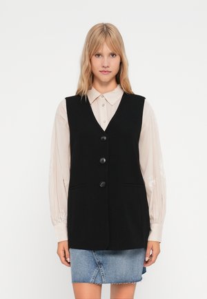 Gilet sans manches noir avec un col en v, cinq boutons et deux poches avant, porté par-dessus une chemise à manches longues de couleur claire et une jupe en denim.