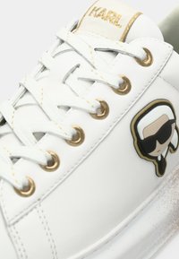 Biała sportowa buta ze złotymi elementami, z teksturowaną powierzchnią, płaskimi sznurowadłami i rysunkowym logo po boku.