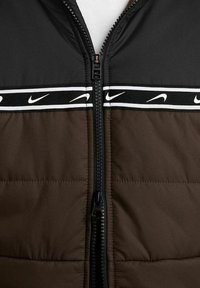 Giacca a vento marrone con parte superiore nera, dotata di una cerniera centrale e del logo Nike bianco su una banda nera che attraversa il petto.