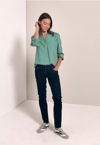 Ljusgrön knappradad skjorta med uppvikta ärmar, tillsammans med mörka jeans och grå sneakers med gröna accenter, stående mot en neutral bakgrund.