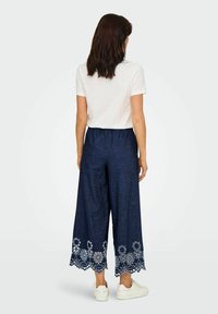 Donkerblauwe, wijde broek met witte geborduurde bloemenpatronen aan de zoom. Elastische tailleband en een vlakke textuur. Draagt een wit T-shirt.