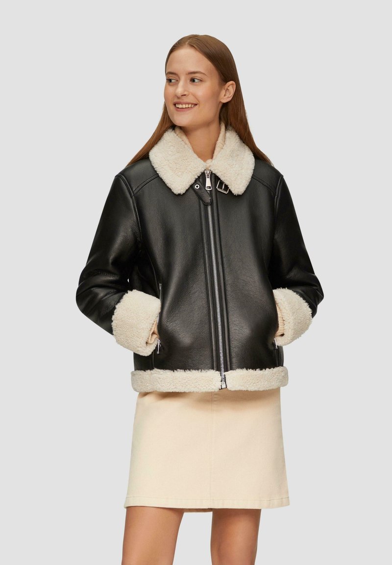 Czarna skórzana kurtka z kremowym kołnierzem i mankietami z baranka, z zamkiem błyskawicznym z przodu i bocznymi kieszeniami. Noszona na beżowej spódnicy.