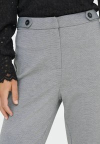 Femme portant un pantalon gris ajusté avec des onglets boutonnés à la taille et un top en dentelle noire à manches longues, la main reposant sur le côté.