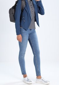 Marinblå blazer med dragkedjedetaljer, randig polo, ljusblå skinny jeans och silverfärgade slip-on skor. Svart quiltad ryggsäck bärs på ena axeln.