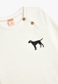 Ecru geribbelde babyshirt met twee houten knopen op de schouder. Voorzien van een zwarte geborduurde honden-silhouet aan de voorkant.