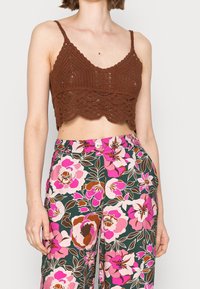 Hnědý háčkovaný crop top s vlnkovaným lemem, kombinovaný s kalhotami s květinovým vzorem, které mají růžové a hnědé květy na tmavě zeleném pozadí.