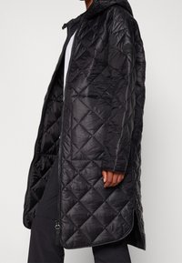 Abrigo acolchado negro con capucha, que presenta una cremallera completa, bolsillos laterales y un patrón de diamantes en la tela. Textura suave y brillante.