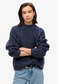 Suéter de punto azul marino con un patrón de trenzado, cuello y puños acanalados, corte oversize, combinado con jeans azul claro y un cinturón negro.