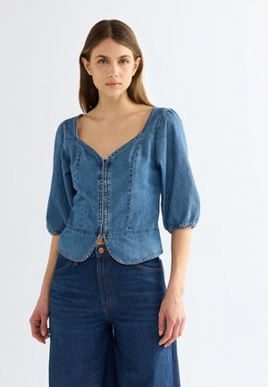 Femme portant un chemisier en denim bleu avec des manches bouffantes et un jean large taille haute bleu foncé, debout devant un fond blanc.