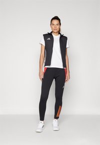 Černá sportovní vesta na zip, bílý tričko, černé legíny s červeným bočním panelem a oranžovými pruhy, doplněné o bílé tenisky.