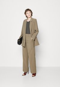 Blazer bej croit cu pantaloni largi asortați și un top gri închis. Modelul ține o poșetă neagră și poartă pantofi cu toc deschisi maro.