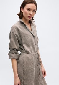 Νεαρή γυναίκα με φόρεμα κουμπωτό σε απόχρωση ταουπέ με λευκές ριγέ λεπτομέρειες, με μανίκια που έχουν τυλιχτεί και ζώνη στη μέση δεμένη, στέκεται μπροστά σε ένα ομοιόμορφο φόντο.