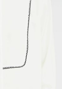 White blouse with a black scalloped trim along the edge. The fabric appears smooth and lightweight with a simple design and no visible patterns.